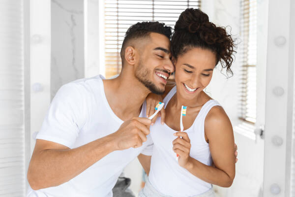 The couple is brushing their teeth and laughing together in the bathroom of their home.