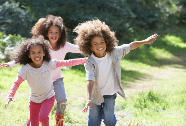 Three happy children with curly hair running and smiling outdoors in a grassy field on a sunny day.