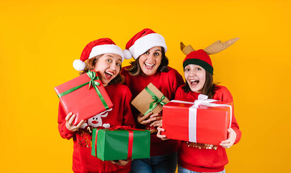 Three women in red sweaters and Santa hats smile while holding gifts in front of a bright yellow background.