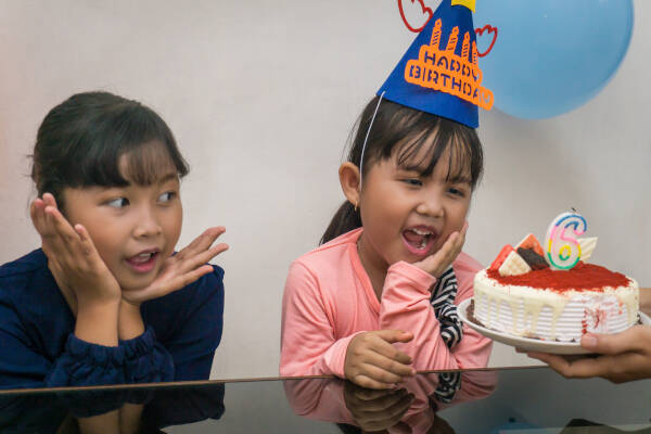 Two excited young girls wearing birthday hats and looking at a cake with a number six candle.