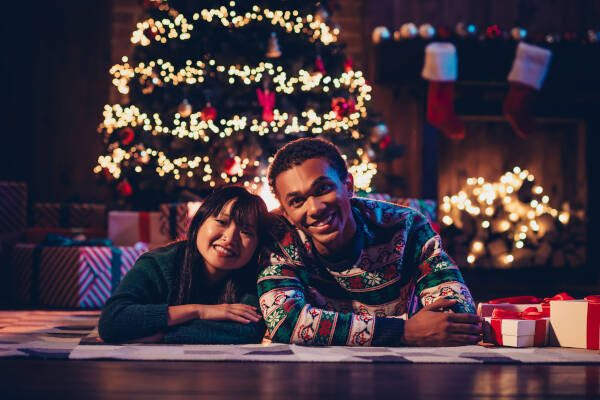 Two friends lying in front of a Christmas tree with presents and lights, smiling at the camera.