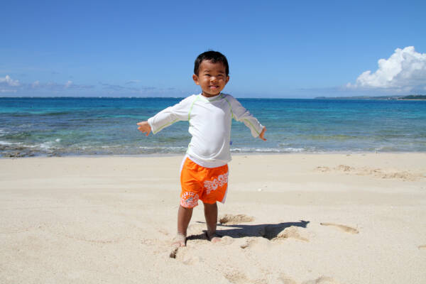 Two-year-old boy stands on a beautiful sunny beach with his arms open, smiling.