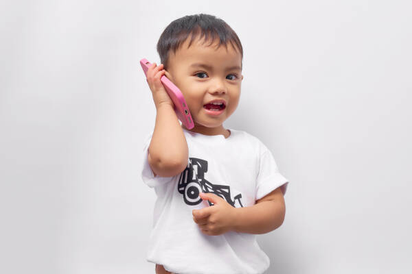 Two-year-old boy wearing a white t-shirt talking on a mobile phone with a friend against a white backdrop.