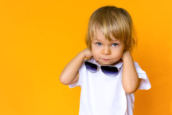 Two-year-old child with blond hair wearing a white t-shirt and sunglasses, set against a yellow background.