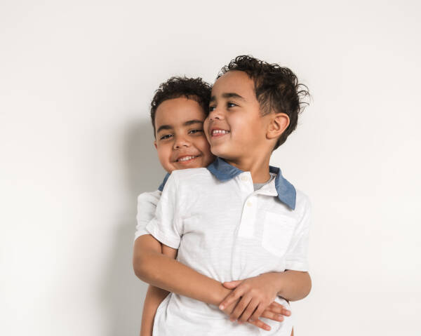 Two young boys smiling and hugging each other while standing against a white background.