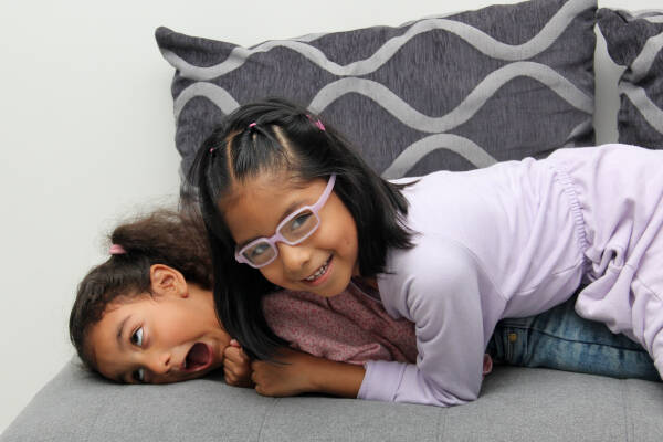 Two young girls playfully lie on a couch, one smiling with glasses, and the other making a silly face.