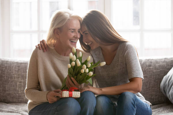 Young adult female child lovingly congratulates excited elderly mother with a gift and flowers on Mother's Day at home.