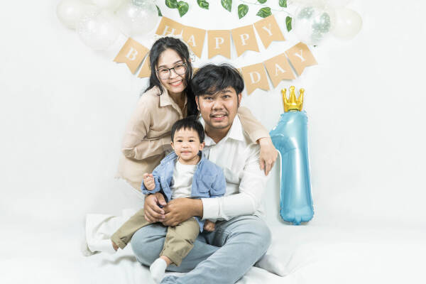 Young couple sits on the floor with their 1-year-old son on his birthday, all smiling. Happy birthday banner and '1' balloon.