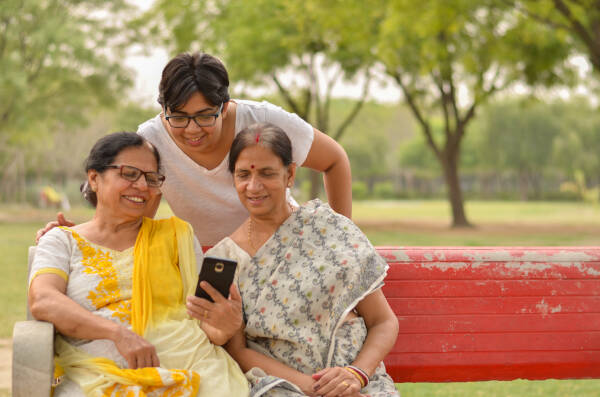 Young girl stands behind park bench with her mother and mother-in-law. They are all looking at photos on a phone and smiling