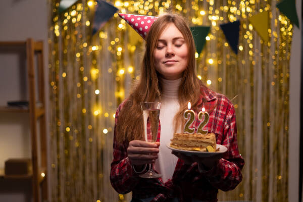 Young woman stands at a party with her eyes closed, making a wish, holding a cake with 22 candles and a glass of champagne.