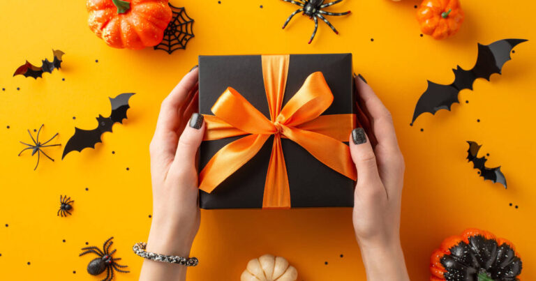 A pair of hands, with black nail polish, holds a black gift box wrapped in an orange ribbon, set against a vibrant orange background decorated with pumpkins, spiders, bats, and spider webs.