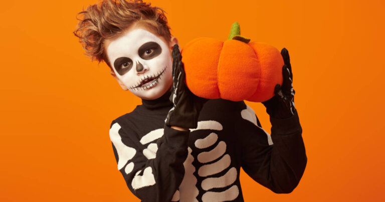 A young boy dressed in a black skeleton costume, his face is painted like a skull, and he is holding a large plush pumpkin with both hands against an orange background.