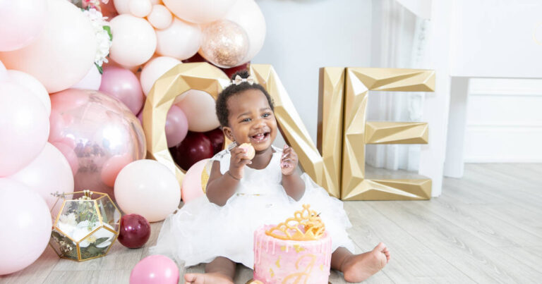 Happy 1-year-old girl smiles at cake smash, ONE sign and balloons in the background.