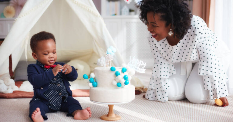 Smartly dressed baby boy celebrates his first birthday with cake at home, mother smiling beside him.