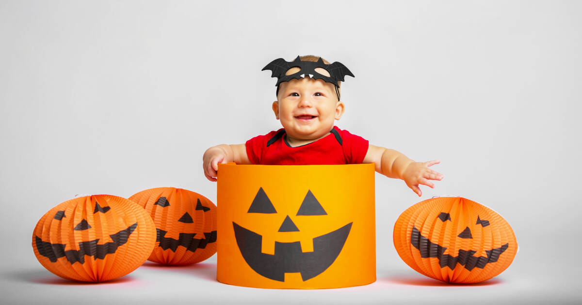 A baby dressed in a red outfit and wearing a bat-shaped headband smiles while sitting inside a large orange jack-o'-lantern prop. Surrounding the baby are orange pumpkin decorations.