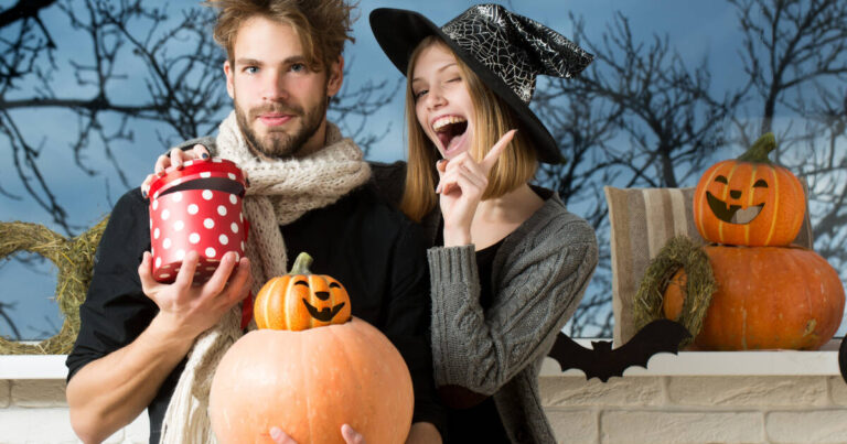 A young couple celebrates Halloween. The man holds a red polka-dotted gift box and a pumpkin, while the woman in a witch hat smiles playfully, surrounded by festive decor.