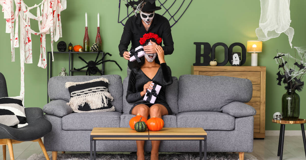 A man in a skeleton costume surprises a woman with a gift box while covering her eyes. They sit in a room decorated with pumpkins, spider webs, and Halloween decor.