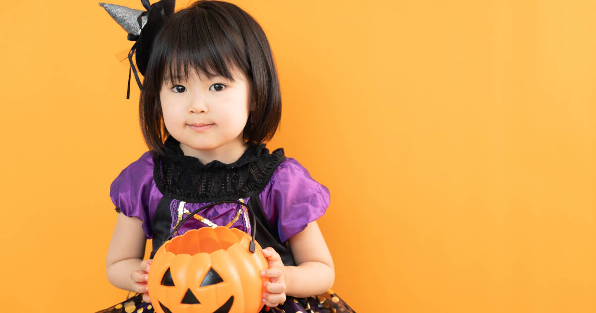 A young girl dressed in a purple Halloween costume with a small witch hat holds a jack-o'-lantern bucket. She stands against an orange background, ready for trick-or-treating.