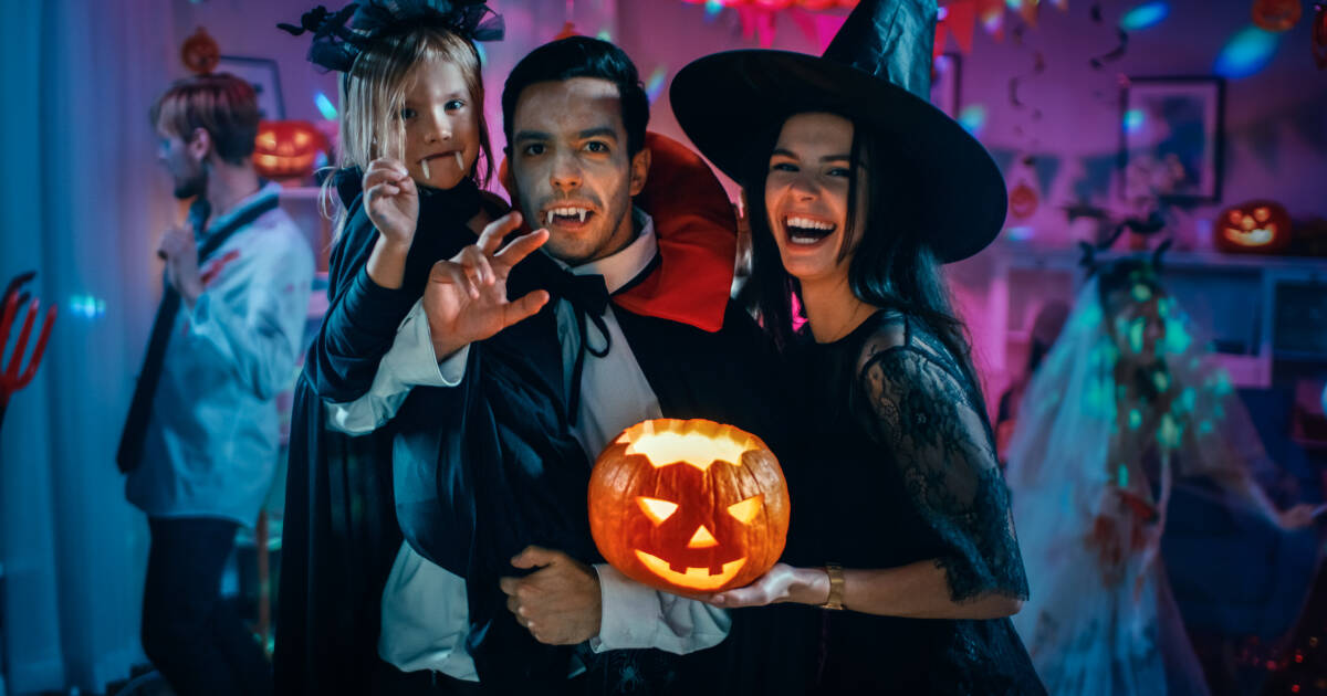 A family dressed in Halloween costumes smiles and poses at a festive party. The father, dressed as a vampire, holds a carved jack-o'-lantern, while the mother, in a witch costume, laughs. A child, also in a vampire outfit, playfully joins in the fun.