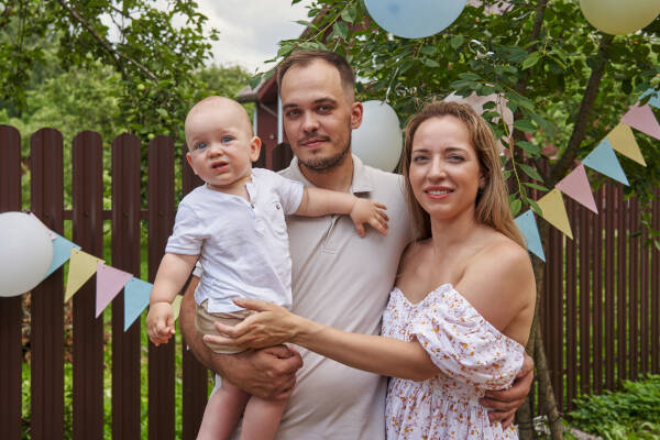 uncle and aunt holding a baby, smiling in front of a decorated backyard with balloons and banners.