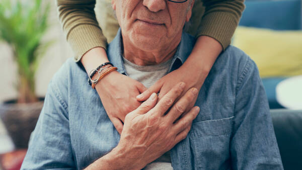 A close-up of an elderly man wearing a denim shirt, with hands wrapped gently around him in a comforting gesture, set in a cozy indoor environment.