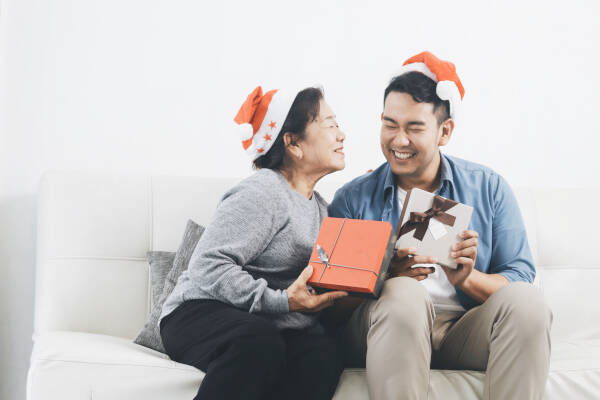 A son and his mother exchanging gifts on the couch, both wearing Santa hats and sharing a joyful moment, as they smile and enjoy the Christmas spirit.