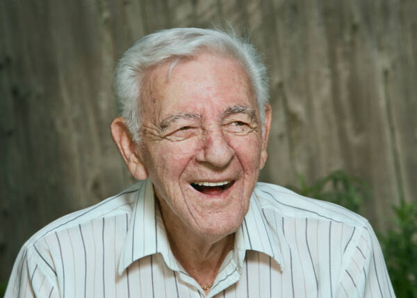 An elderly man with white hair, wearing a striped shirt, smiling joyfully against a blurred wooden background.