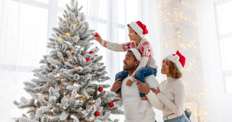 A joyful family decorating a snow-covered Christmas tree together, with a young child on the father's shoulders adding ornaments, all wearing festive Santa hats.