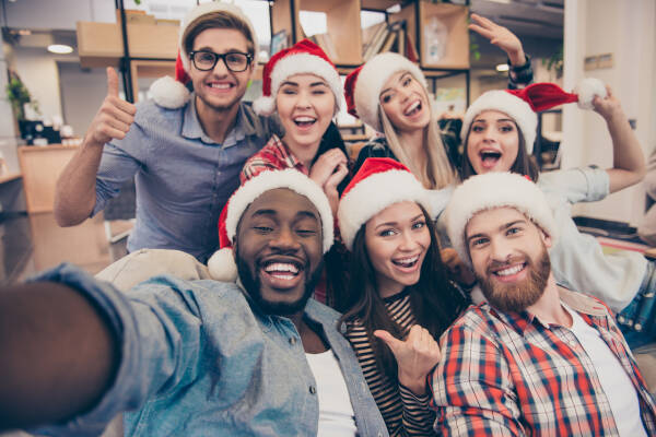 Diverse group of young professionals in Santa hats taking a cheerful selfie at a festive holiday gathering, enjoying the season's spirit.