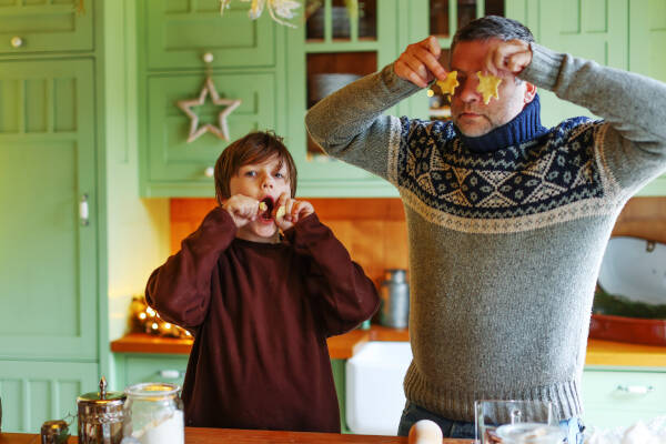 Father and son making funny faces while holding Christmas cookies in front of their eyes, enjoying a playful holiday moment together in a festive kitchen.