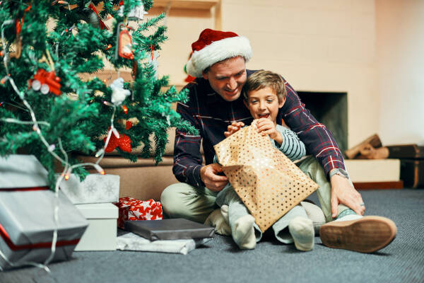 Father and young son excitedly unwrapping a Christmas present together near a decorated Christmas tree, sharing a joyful holiday moment in a cozy living room.