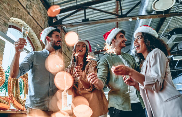 Group of colleagues wearing Santa hats celebrating at a holiday party with sparklers and champagne, sharing joyful moments together.