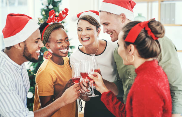 Group of friends in festive holiday attire, toasting with glasses of red wine and laughing, creating warm and joyful holiday memories together.