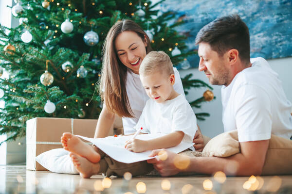 Mom, dad, and young child sitting on the floor by a Christmas tree, as the child excitedly draws on paper, surrounded by holiday decorations and warm lighting.