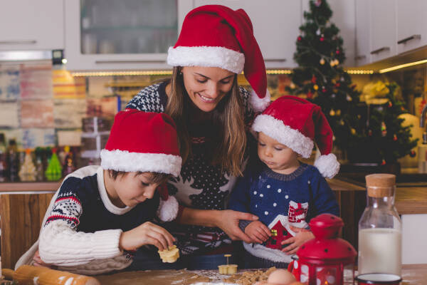 Mother and her two young children baking cookies in a holiday-themed kitchen, all wearing Santa hats and sharing a warm family Christmas activity.