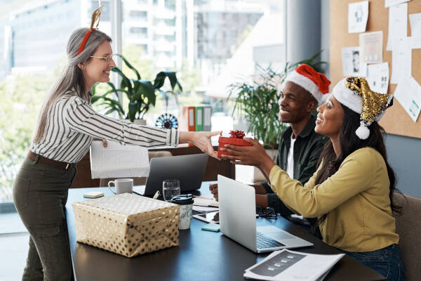 Office team wearing Christmas hats gathered around a table with gifts, celebrating the holiday season in a modern workspace.