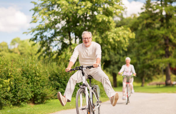 A cheerful older man riding a bicycle with his legs in the air, followed by a smiling older woman cycling on a sunny park path.