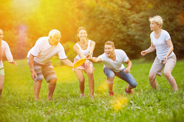 A group of people of mixed ages laughing and reaching for a frisbee while playing outdoors on a sunny day in a grassy field.