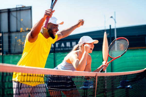 A man and a woman playing tennis on an outdoor court, celebrating a successful point with raised arms and wide smiles.