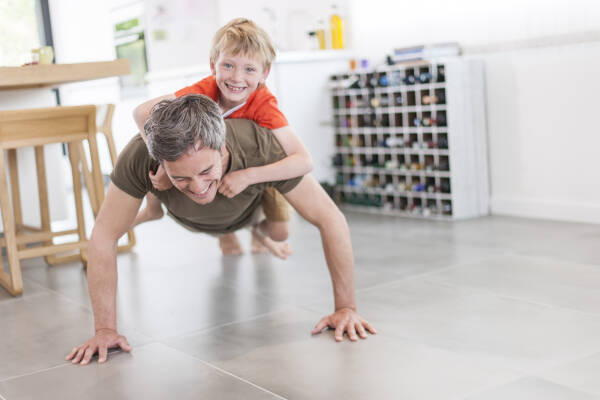 A young boy riding on a man's back as he does push-ups inside a bright, modern home, both laughing joyfully.