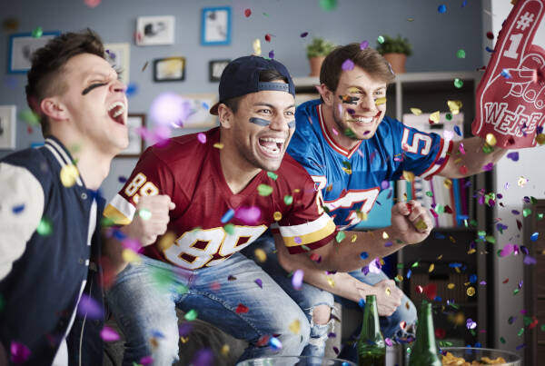 Three excited young men wearing sports jerseys, cheering enthusiastically while watching a game indoors, surrounded by confetti.