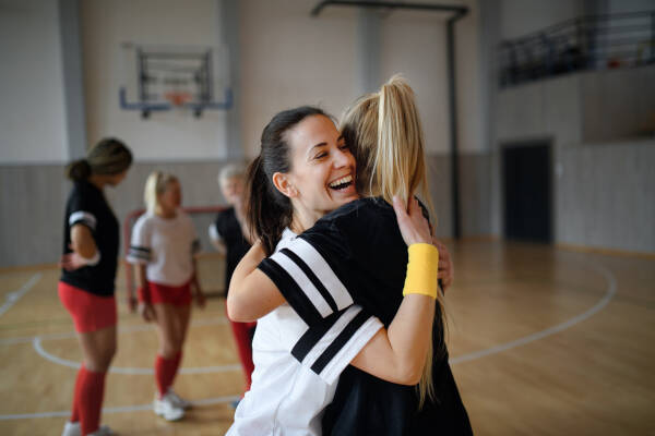 Two female volleyball players hugging in celebration on an indoor court while teammates look on in the background.