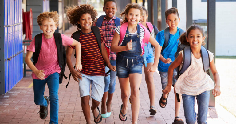 A group of smiling children, wearing backpacks, running enthusiastically down a school corridor lined with lockers.