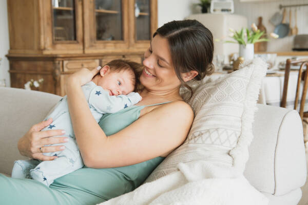 A smiling woman sitting on a cozy couch holds a baby dressed in a light blue onesie with star patterns.