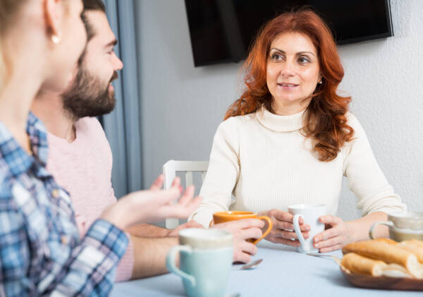 Three people sitting at a table enjoying coffee and conversation; a red-haired woman in a white turtleneck is engaged in discussion with two younger individuals.