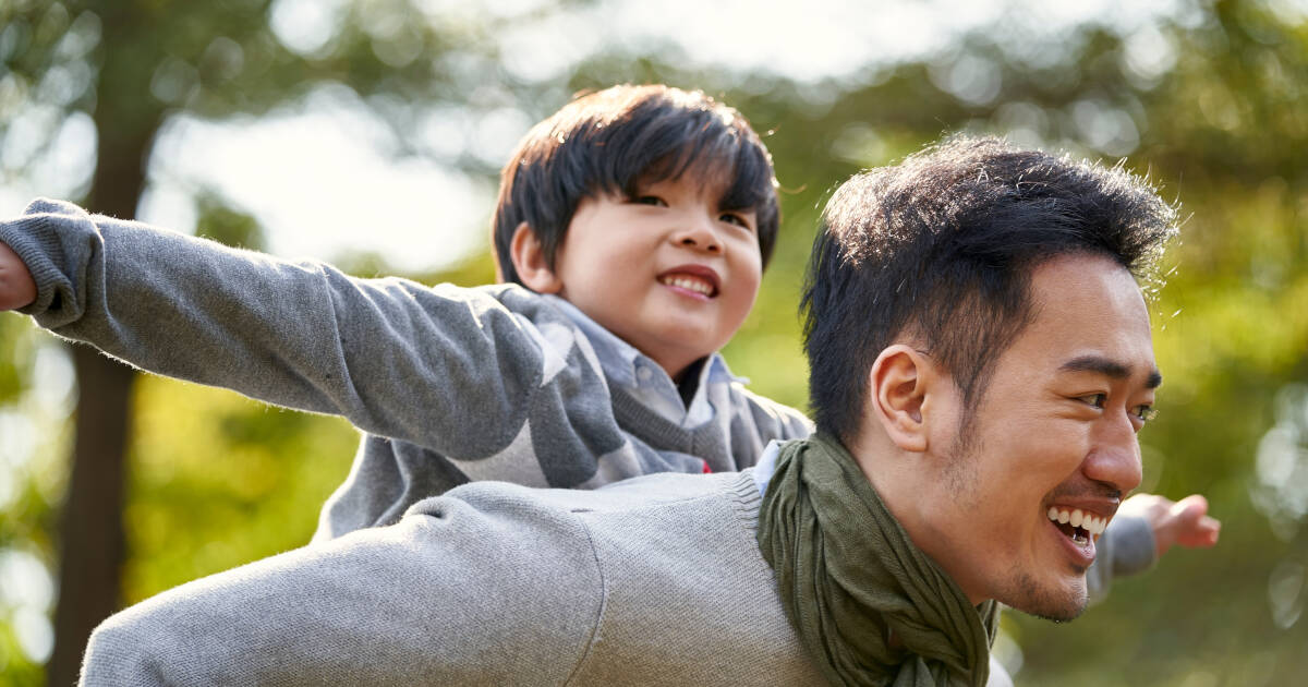 Young asian father carrying son on back having fun enjoying nature outdoors in park.