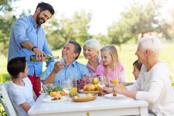 Family having a cheerful outdoor meal, with one man pouring a drink for an older man.