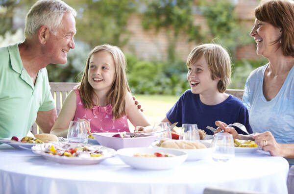 Grandparents and two children sitting at an outdoor table, smiling and talking over a meal.
