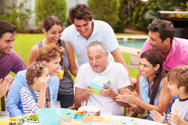 Large family gathered around an elderly man opening a birthday gift at an outdoor table.