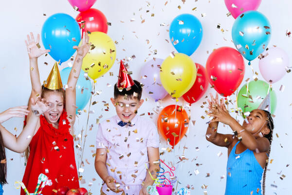 Three kids in party hats celebrating with confetti and balloons at a birthday party.